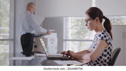Side view of caucasian businesswoman sitting at desk in office using computer. Young female manager working on laptop in modern office with colleague using copier on background