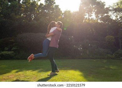 Side View Of A Caucasian Couple In The Garden, Embracing On A Sunny Day, The Man Lifting The Woman Off Her Feet, Backlit With Lens Flare 