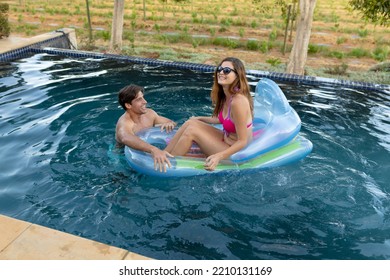 Side view of a Caucasian couple in a swimming pool, the man standing and holding onto an inflatable pool lounger and the woman sitting on it wearing sunglasses and smiling - Powered by Shutterstock