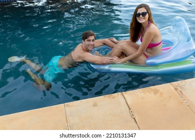 Side view of a Caucasian couple in a swimming pool, the man standing and holding onto an inflatable pool lounger and the woman sitting on it wearing sunglasses, both smiling to camera - Powered by Shutterstock