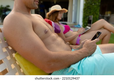 Side View Of A Caucasian Couple Wearing Beachwear Sitting On Chairs Sunbathing In A Garden, The Woman Wearing A Sun Hat And Using A Laptop, The Man Using A Smartphone