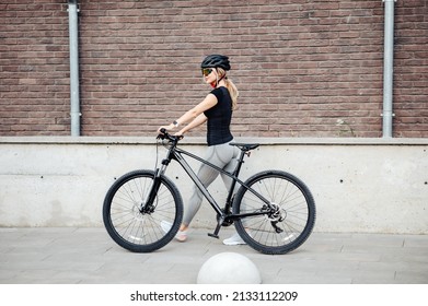 Side View Of Caucasian Female Cyclist In Activewear, Helmet And Glasses Walking With Black Bike On City Street. Relaxation After Workout.