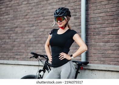 Side View Of Caucasian Female Cyclist In Activewear, Helmet And Glasses Standing With Black Bike On City Street. Relaxation After Workout.