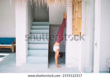 Side view of caucasian girl practicing yoga in Wall-assisted handstand pose at home. Concept of healthy lifestyle. Young beautiful flexibility woman wearing sportswear and barefoot. Sunny daytime