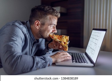 Side view of Caucasian hardworking architect eating pizza and using laptop while sitting in office late at night. - Powered by Shutterstock