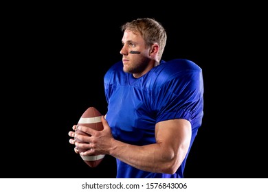 Side View Of A Caucasian Male American Football Player Wearing A Team Uniform, Pads And Eye Black Under His Eyes, Holding A Football In His Hands