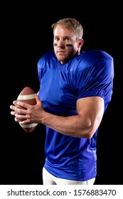 Side View Of A Caucasian Male American Football Player Wearing A Team Uniform, Pads And Eye Black Under His Eyes, Holding A Football In His Hands And Turning To Camera. Vertical Shot