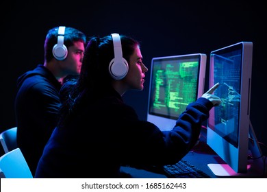 Side view of a Caucasian man and a woman working late in an office in the evening together, using computers, programming pointing and looking at computer screens, wearing headphones in a darkly lit