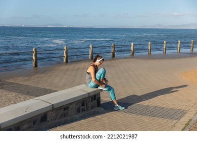 Side View Of A Caucasian Mature Middle Aged Woman Working Out On A Promenade On A Sunny Day With Blue Sky, Sitting, Tying Her Shoe Laces