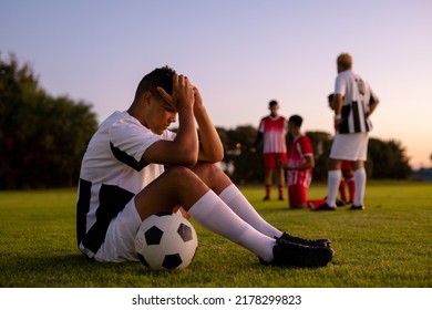 Side view of caucasian sad male player with head in hands and ball sitting on field at sunset. Copy space, playground, frustration, loss, stress, unaltered, soccer, competition and sport concept. - Powered by Shutterstock