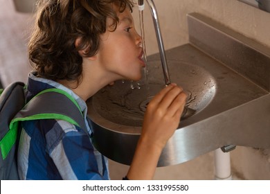 Side View Of A Caucasian Schoolboy Drinking Water From Tap In The Corridor At School