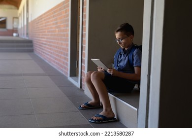 Side View Of A Caucasian Schoolboy Wearing Glasses, Shorts, Flip-flops And A Rucksack Sitting On A Step By A Wall In The Schoolyard At Elementary School Using A Tablet Computer