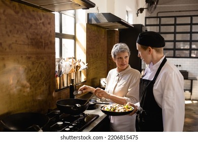 Side view of a Caucasian Senior woman using a wok and a Caucasian female chef holding a dish of chopped vegetables talking during a cookery class in a restaurant kitchen - Powered by Shutterstock