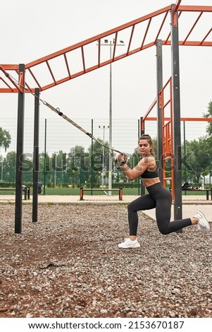 Side view of caucasian sportswoman doing lunges exercise with suspension straps on sports ground. Focused adult woman wearing sportswear. Modern healthy and sports lifestyle. Cloudy daytime