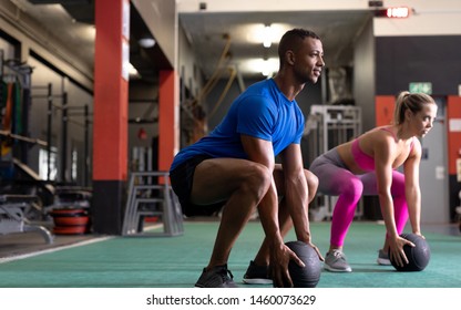 Side view of an Caucasian woman and an African-American man lifting ball weights while doing squats inside a room at a sports centre