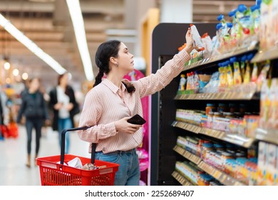 Side view of Caucasian woman choosing products in grocery store. Student reaches hand to top shelf holds cellphone and cart. Shelves with food in background. Concept of shopping and consumerism. - Powered by Shutterstock