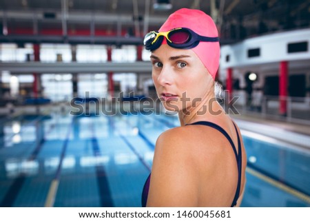 Side view of a Caucasian woman wearing a swimsuit and a pink swimming cap with goggles looking at the camera while standing by an olympic sized pool inside a stadium