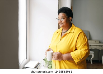 Side view of charming elderly plus size african american woman in yellow shirt holding white cup of hot drink staring through window at noisy neighborhood with smile, having rest and relaxing - Powered by Shutterstock