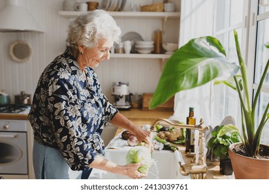 Side view of charming kind caucasian grandma washing vegetables from her garden under tap water in kitchen to cook delicious tasty dinner for her grandchildren, friends and big family - Powered by Shutterstock