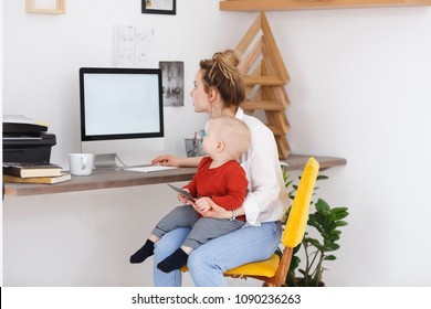 Side view of charming young stylish mother holding in hands and a cute baby sitting at the desk on white wall background. Place for advertising