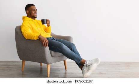 Side View Of Cheerful African American Guy Drinking Coffee Sitting In Chair Over Gray Background. Black Man Enjoying Weekend Relaxing With Hot Drink Indoors, Looking Aside. Panorama