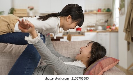 Side View Cheerful Asian Mother Lying On Sofa And Holding Up Daughter On Legs At Home. The Girl Open Arms Like She’s Flying