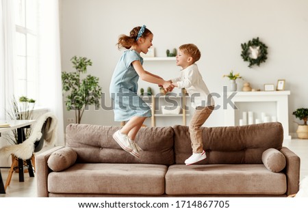 Side view of cheerful boy and girl holding hands and jumping on couch while having fun at home together
