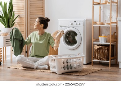Side view of cheerful brunette woman holding clothes near basket in laundry room - Shutterstock ID 2265425297