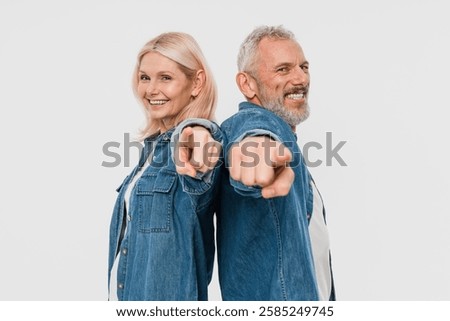 Side view of cheerful couple two friends elderly gray-haired man blonde woman in denim clothes standing back to back pointing index finger on camera isolated on white background studio portrait