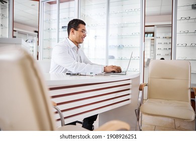 Side view of a cheerful dark-haired young optician in round eyeglasses typing on his laptop - Powered by Shutterstock