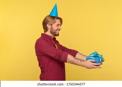 Side view, cheerful delighted guy in funny party cone holding wrapped present, giving gift box greeting happy birthday, congratulating on anniversary. studio shot isolated on yellow background