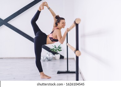 Side view of cheerful female ballet dancer joyfully stretching at wooden barre holding foot above head in light dancing studio