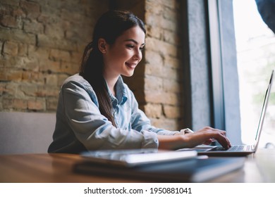 Side View Of Cheerful Female Remote Worker With Toothy Smile Sitting At Table Near Window And Using Laptop For Work