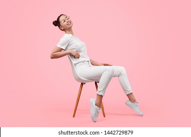 Side view of cheerful female teenager in stylish white outfit smiling and leaning back while sitting on chair against pink background - Powered by Shutterstock