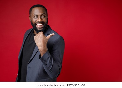 Side View Of Cheerful Funny Young African American Business Man 20s Wearing Classic Jacket Suit Standing Pointing Thumb Aside On Mock Up Copy Space Isolated On Bright Red Background Studio Portrait