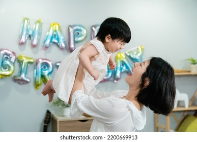 Side View Of A Cheerful Korean Mother Holding Her Lovely Baby Daughter High Up With The Wall Decorated For Birthday At Background.