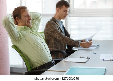 Side View Of Cheerful Man In Green Shirt Relaxing In Chair In Office Building. Male Worker Sitting At Table, Stretching And Laughing During Working Day. Concept Of New Idea And Happiness.