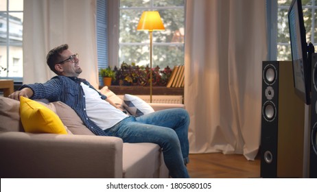 Side View Of Cheerful Man Watch Comedy TV Show Relaxing In Living Room. Happy Handsome Guy Resting On Couch Enjoying Movie And Laughing Staying At Home