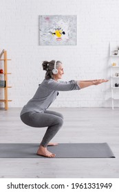 Side View Of Cheerful Middle Aged Woman In Sportwear And Headphones Doing Squat On Mat At Home