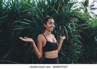 Side view of cheerful Middle Eastern female with raised hands smiling during summer vacations in Indonesia, happy woman in sportive top enjoying leisure time near tropical palm trees in Bali