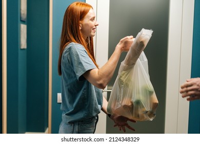 Side view of cheerful redhead female customer receiving groceries bag from unrecognizable delivery man on doorstep at apartment. Cropped shot of courier male delivering food meal to client at home. - Shutterstock ID 2124348329