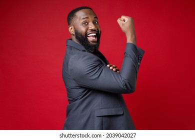 Side View Of Cheerful Strong Young African American Business Man 20s Wearing Classic Jacket Suit Standing Showing Biceps Muscles Looking Camera Isolated On Bright Red Color Background Studio Portrait