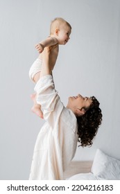 Side View Of Cheerful Woman Lifting Baby Daughter In Bedroom