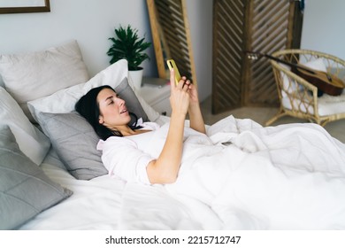 Side View Of Cheerful Young Female Resting On Comfortable Bed While Looking At Screen Of Mobile Phone And Enjoying Free Time In Bedroom