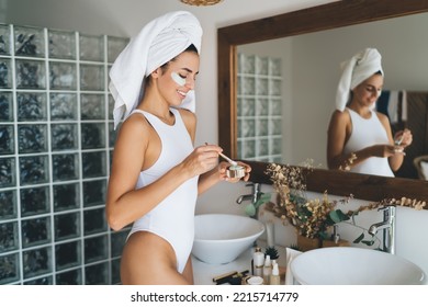 Side View Of Cheerful Young Female In Bodysuit Standing With Brush And Cream In Hands While Preparing To Moist Face Before Makeup