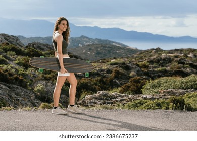 Side view of cheerful young female skateboarder with longboard strolling on walkway while looking away against mountain and sea - Powered by Shutterstock