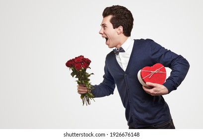 Side View Of Cheerful Young Romantic Guy In Elegant Outfit Carrying Bouquet Of Red Roses And Heart Shaped Gift Box, While Rushing To Date On White Background