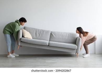 Side View Of Cheery Young Asian Couple Moving Sofa At Home, Replacing Furniture In Living Room, Blank Space. Happy Millennial Spouses Making Rearrangement In Their House Together