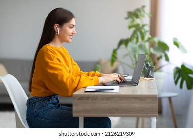 Side view of cheery young woman in earphones using laptop, having online business meeting or educational webinar at home, copy space. Remote communication, work conference, distance lesson concept - Shutterstock ID 2126219309