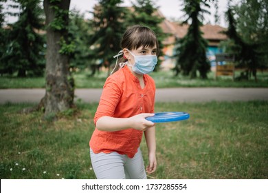 Side View Of Child Holding Flying Disk And Breathing Through Medical Mask. Nice Girl Playing Outside In Front Of Green Trees During The Coronavirus Pandemic